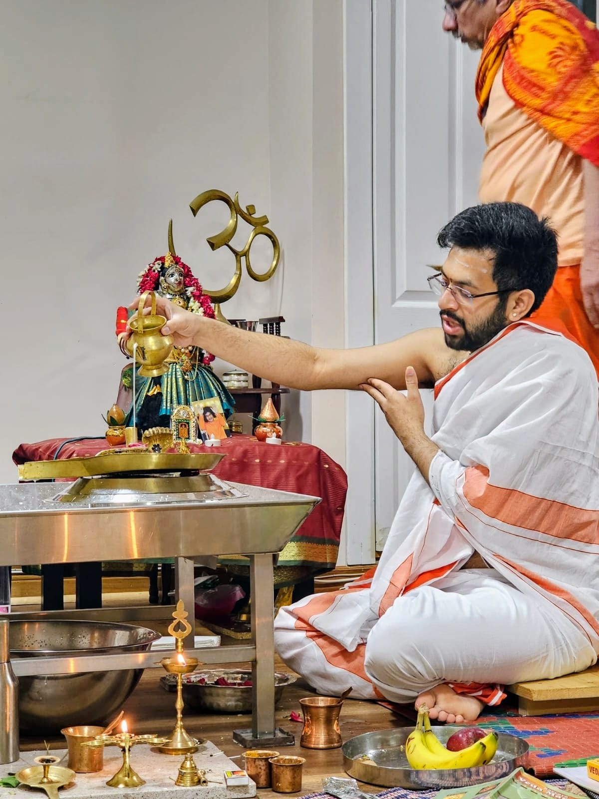 Purohit seated on the floor performing ritual water offerings at a home altar